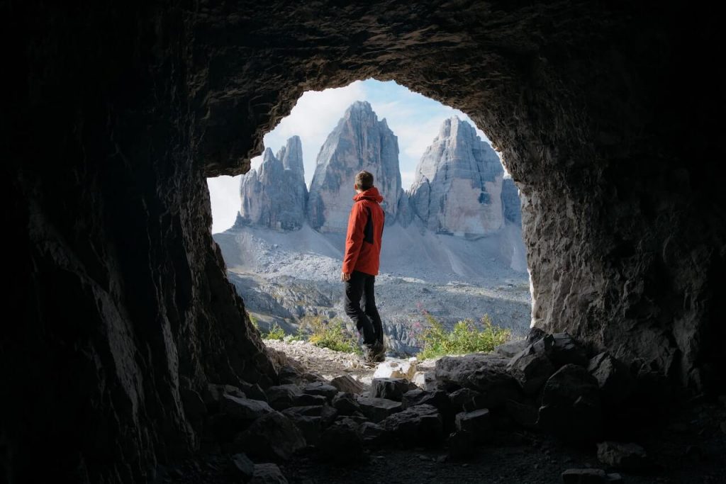 Una imagen de una persona de pie en una montaña, frente a una impresionante vista de las montañas circundantes, con la entrada de una cueva a sus espaldas.